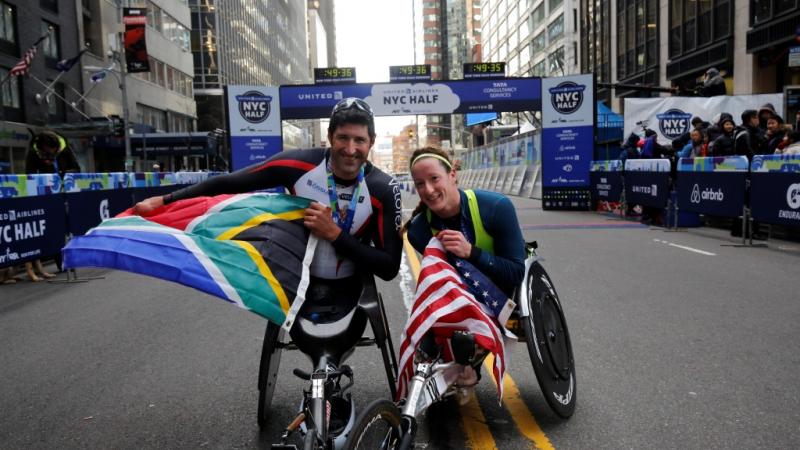 A man and a woman in racing wheelchairs, posing and showing an American and a South African flag