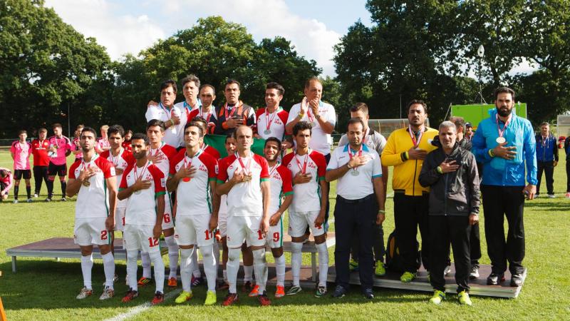 Iranian football 7-a-side team and their staff stand together, put their right hand ver their chests.  