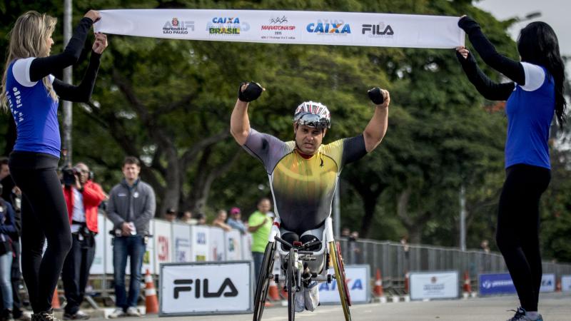 Man in wheelchair raises both arms as he crosses a finishline. 