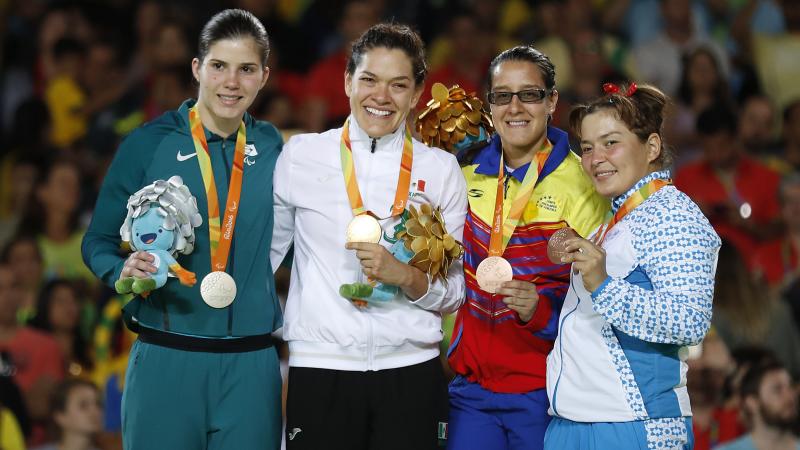 Four women in training suits on a podium, showing medals and mascots