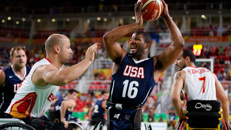 Man in wheelchair holds a basketball in the air away from his defender