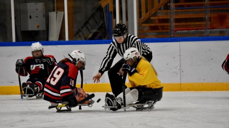 Two female sledge hockey players on the ice, fighting for the puk from the referee