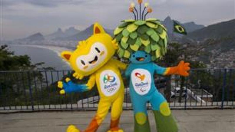 A yellow mascots and blue mascot with green leaves as hair stand on top of Rio's Sugar Loaf mountain.