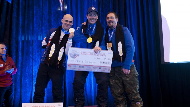 Three men on a podium holding giant checks and their medals