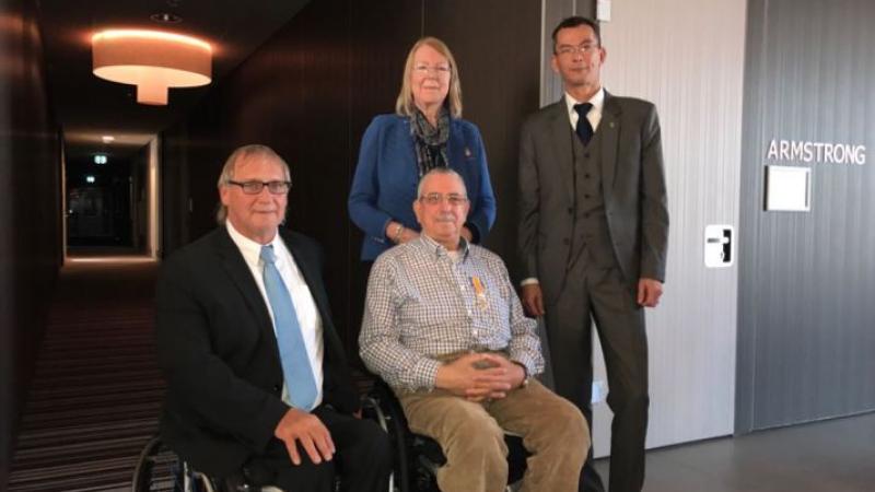 Two men in wheelchairs in suits while two people stand behind them to pose for a photo