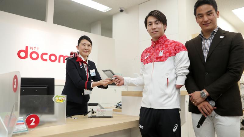 Two Japanese men pose for a photo as a woman hands over a cell phone