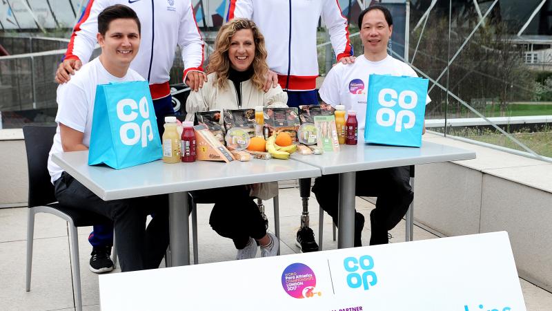 Two men in tracksuits stand behind three people sat at a table.  In the background is an athletics stadium.