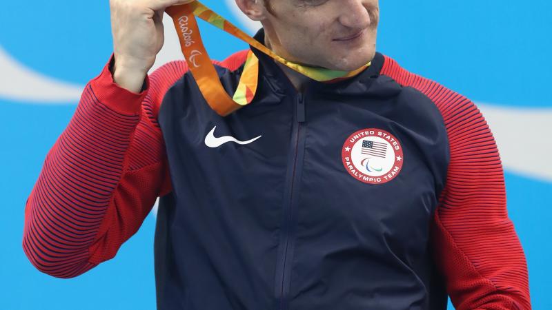 Blind man on podium shakes his gold medal near his ear