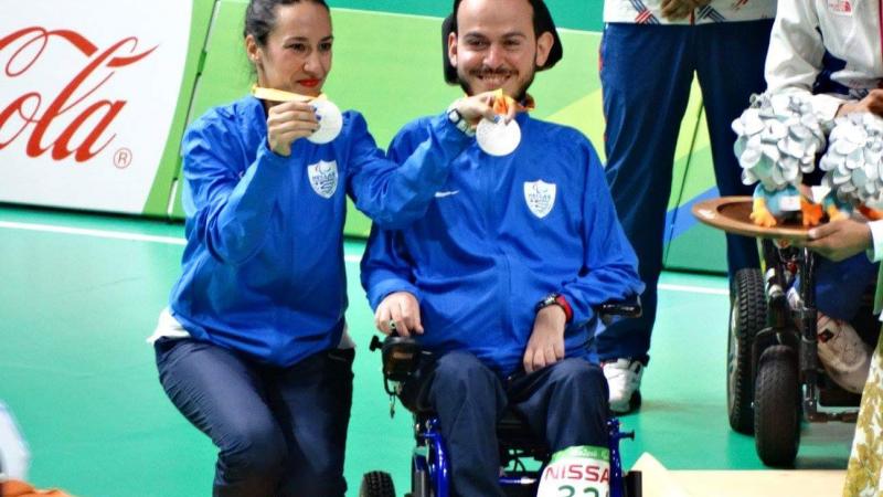 a woman and a man in a wheelchair hold up their silver Paralympic medals