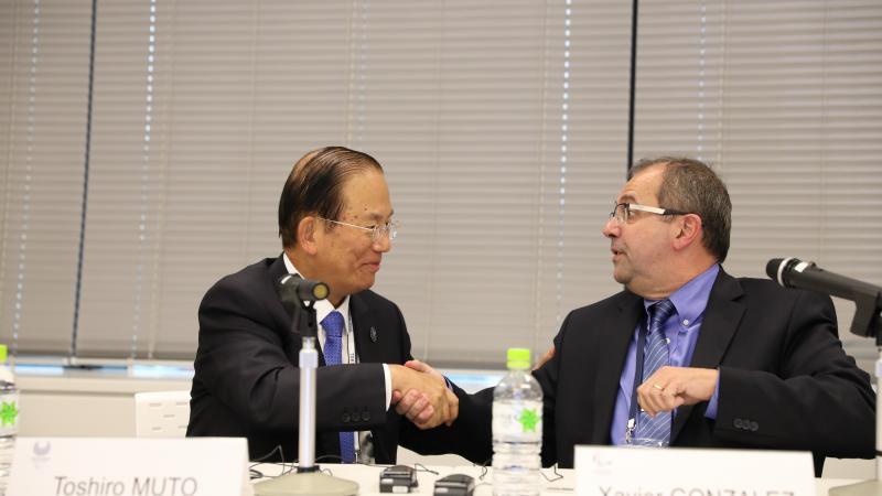 two men shake hands across a table and smile