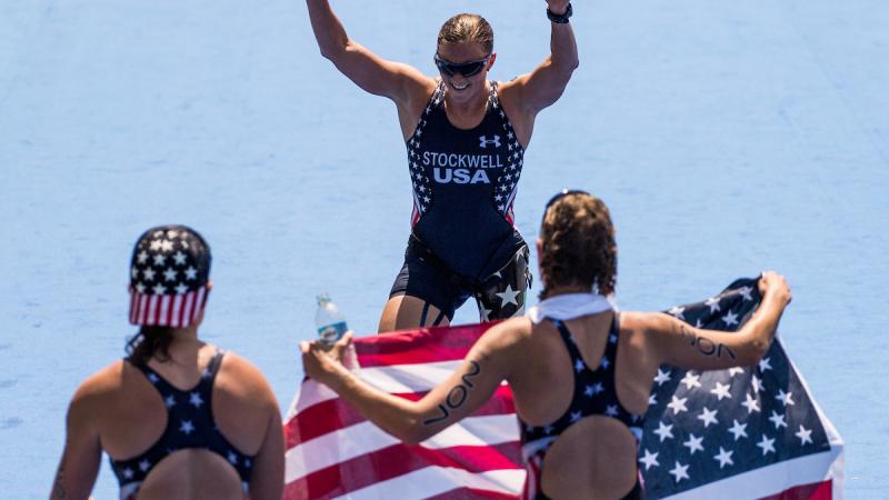 Two women holding the US flag waiting for their teammate to cross the finish line