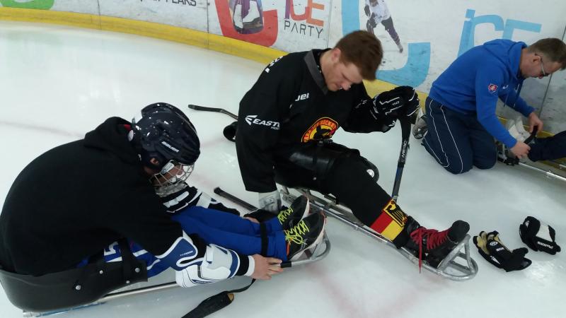 Two men in Para ice hockey sledges, one helping another get in the sledge
