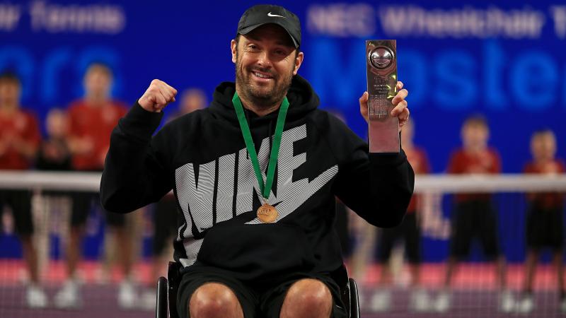 a male wheelchair tennis player holds up a trophy