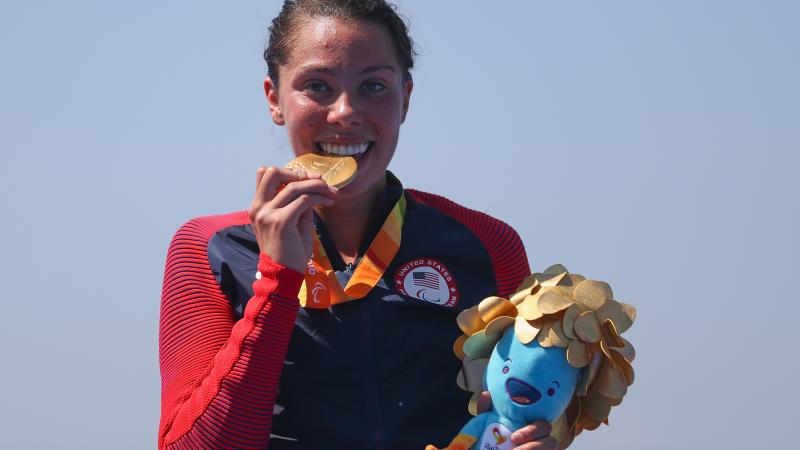 a female Para triathlete stands on the podium and bites her gold medal