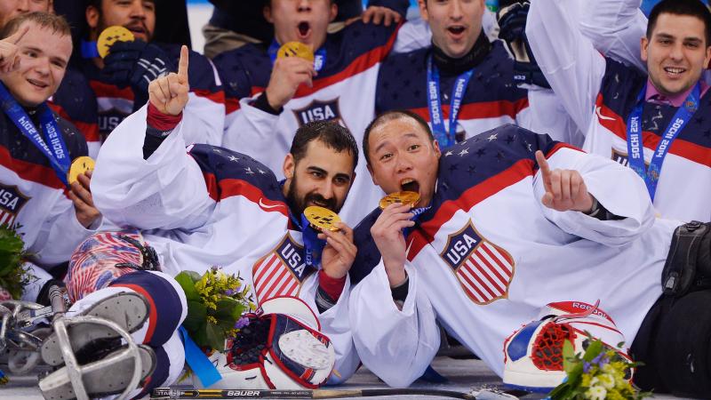 a group of male para ice hockey players lie on the ice and bite their medals in celebration