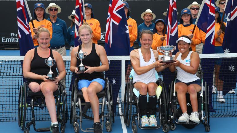 four female wheelchair tennis players hold up their trophies on court