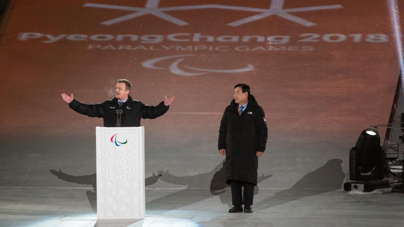 a man spreads his arms wide while speaking at a podium