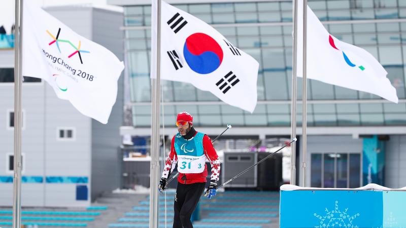 a male Para Nordic skier walks past the Paralympic and South Korean flags