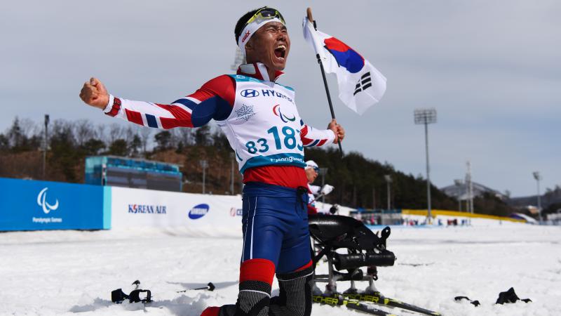 a sit skier celebrates on his knees holding the South Korean flag