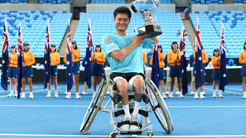 a male wheelchair tennis player lifts a trophy