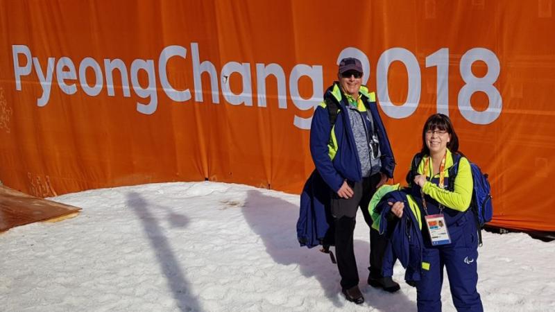 Two winter sport officials pose in front of a PyeongChang 2018 banner