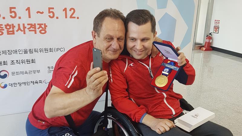 a male Para shooter and his coach hold up his gold medal