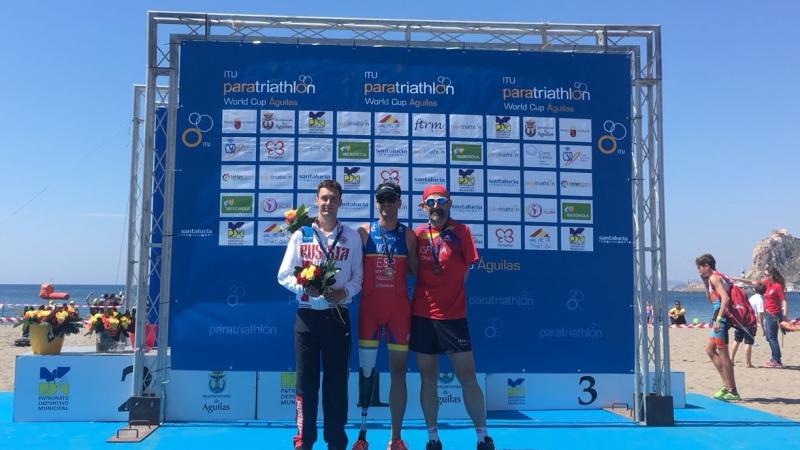 three male Para triathletes on a podium on the beach