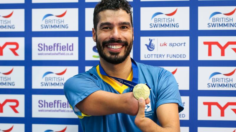 a male Para swimmer smiles while holding up his gold medal