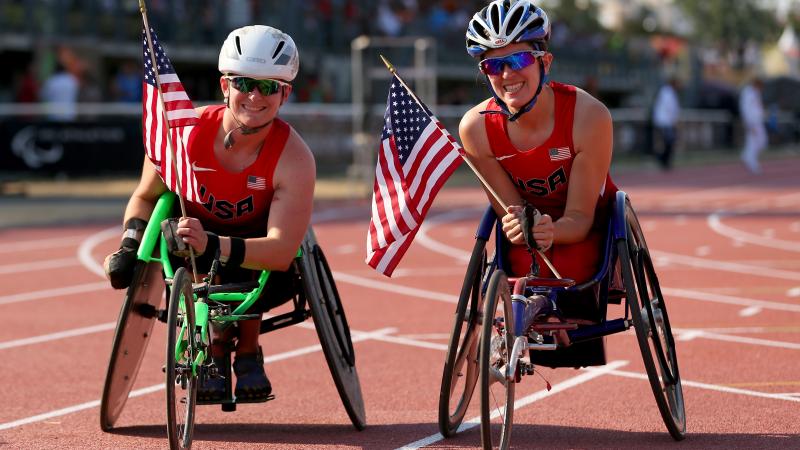 two female wheelchair racers hold up American flags on the track