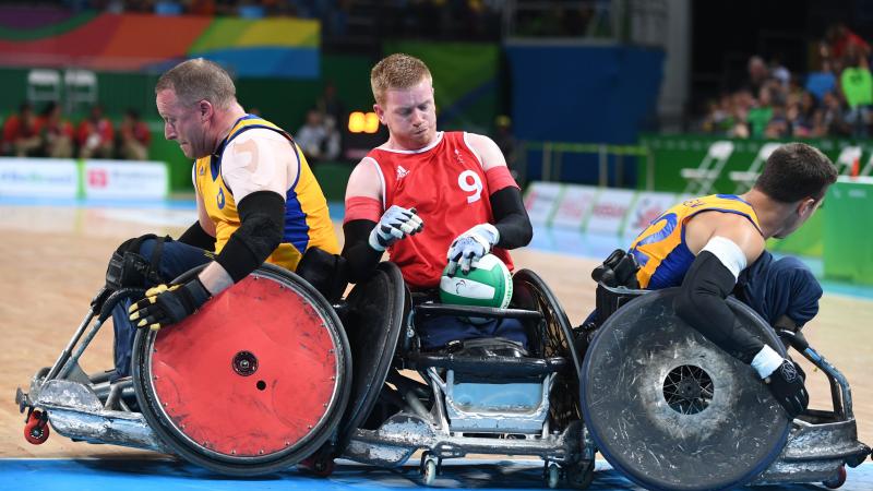 a male wheelchair rugby player holds onto the ball while being challenged by two other players