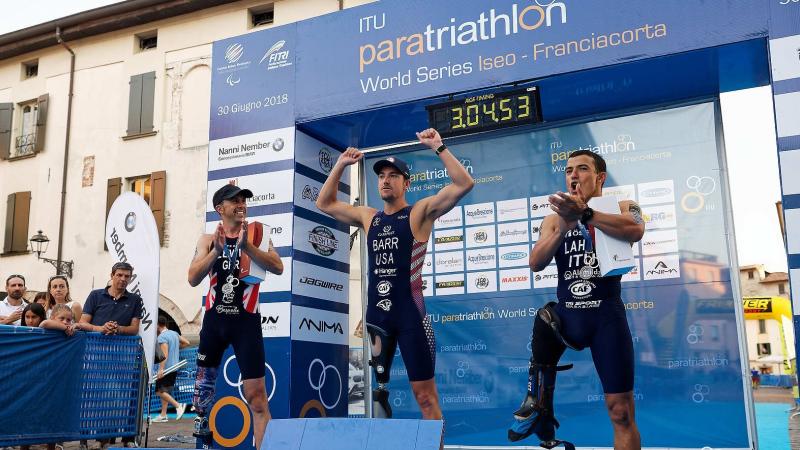 three male Para triathles on a podium with the middle one raising his arms