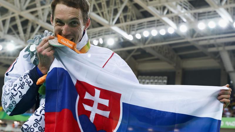 male Para cyclist Jozef Metelka bites a gold medal and holds up a Slovenian flag