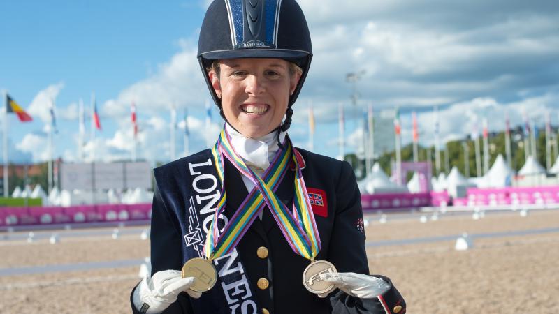 female Para equestrian rider Erin Orford smiles and holds up two gold medals around her neck
