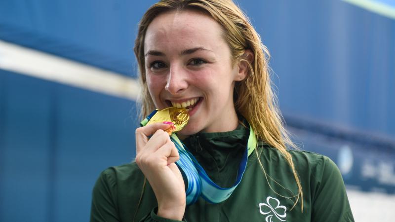 female Para swimmer Ellen Keane smiles and bites her gold medal