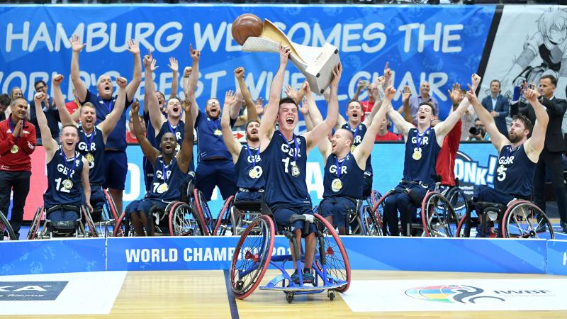 Man in wheelchair hoists basketball trophy with the men's British wheelchair basketball team behind him celerbating 