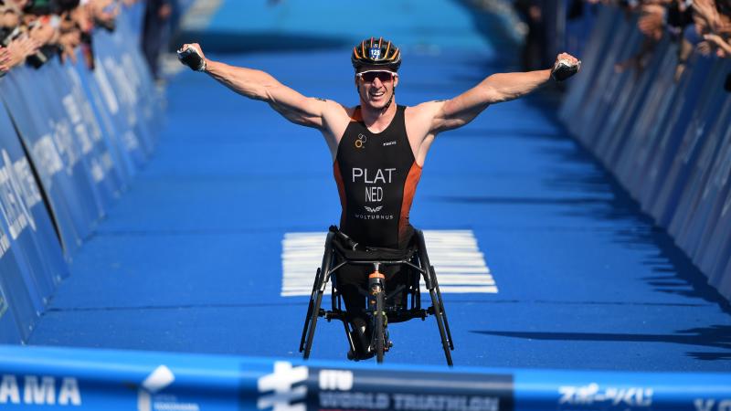 male Para triathlete raises his arms in celebration as he breaks the finish line tape