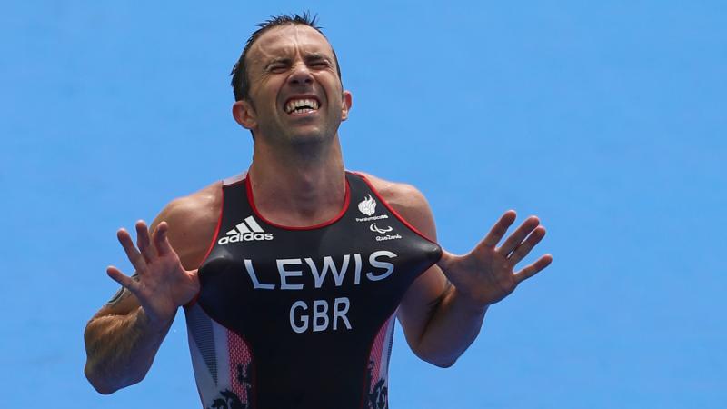 male Para triathlete Andy Lewis points his fingers to the sky as he crosses the finish line