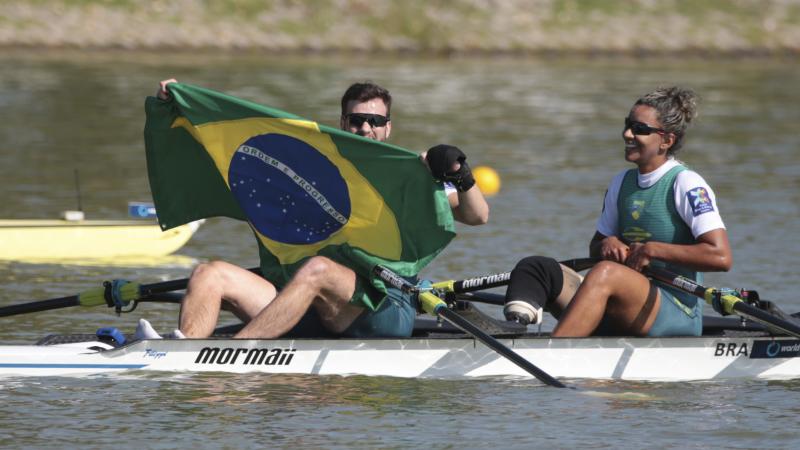 A man and woman in rowing boat celebrate, while man holds up Brazilian flag