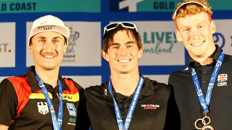 three male Para triathletes with Stefan Daniel in the centre, smiling on the podium with their medals