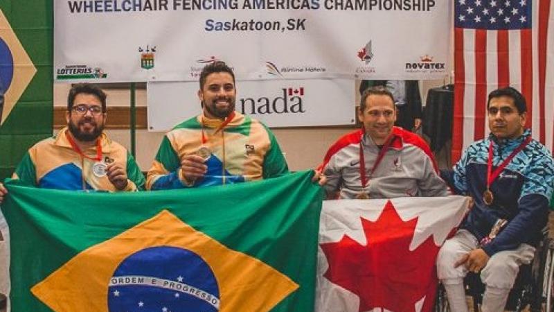 four male wheelchair fencers on the podium including Jovane Guissone holding a Brazilian flag