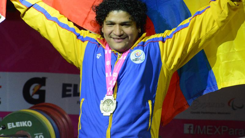 a male powerlifter smiling with his medal round his neck holding up a Colombian flag