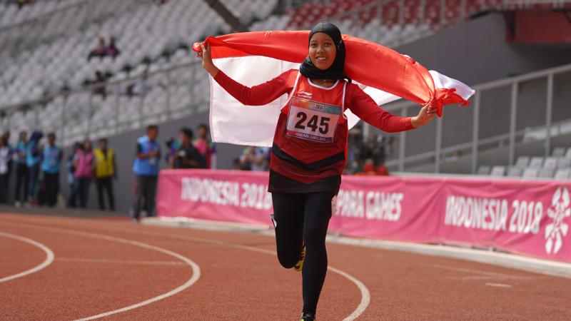 female Para athlete Karisma Tiarani runs down the track holding an Indonesia flag