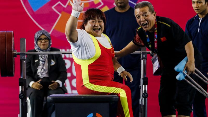 female powerlifter Xu Lili waves and smiles while sitting on the bench