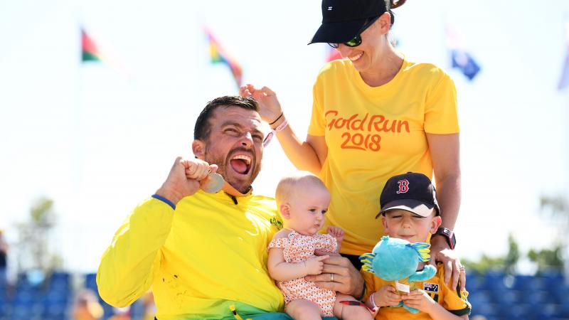 male wheelchair racer Kurt Fearnley holds up his medal alongside his wife and holding his child on his lap