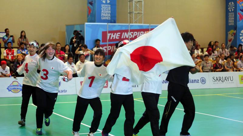 Japanese female goalball players run across the court holding a Japan  flag
