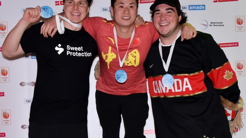 male Para snowboarder Sun Qi stands on the top of the podium with his arms around two other snowboarders