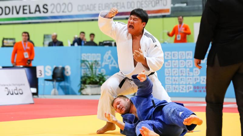 A man in a white kimono celebrates while other man in blue kimono lies on the mat