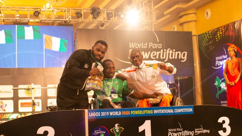 three male powerlifters, with Mufutau Rasaq in the middles, link arms on the podium