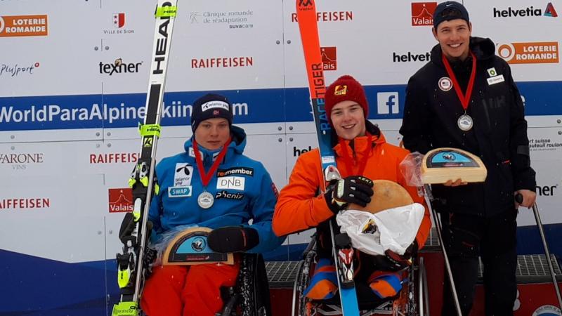 three Para alpine skiers holding up wheels of cheese on the podium