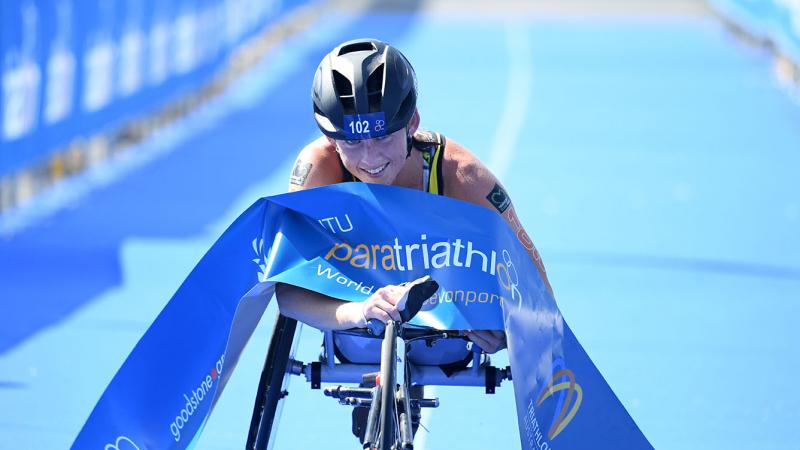 Australian woman in racing wheelchair crosses the finishline tape at a triathlon event